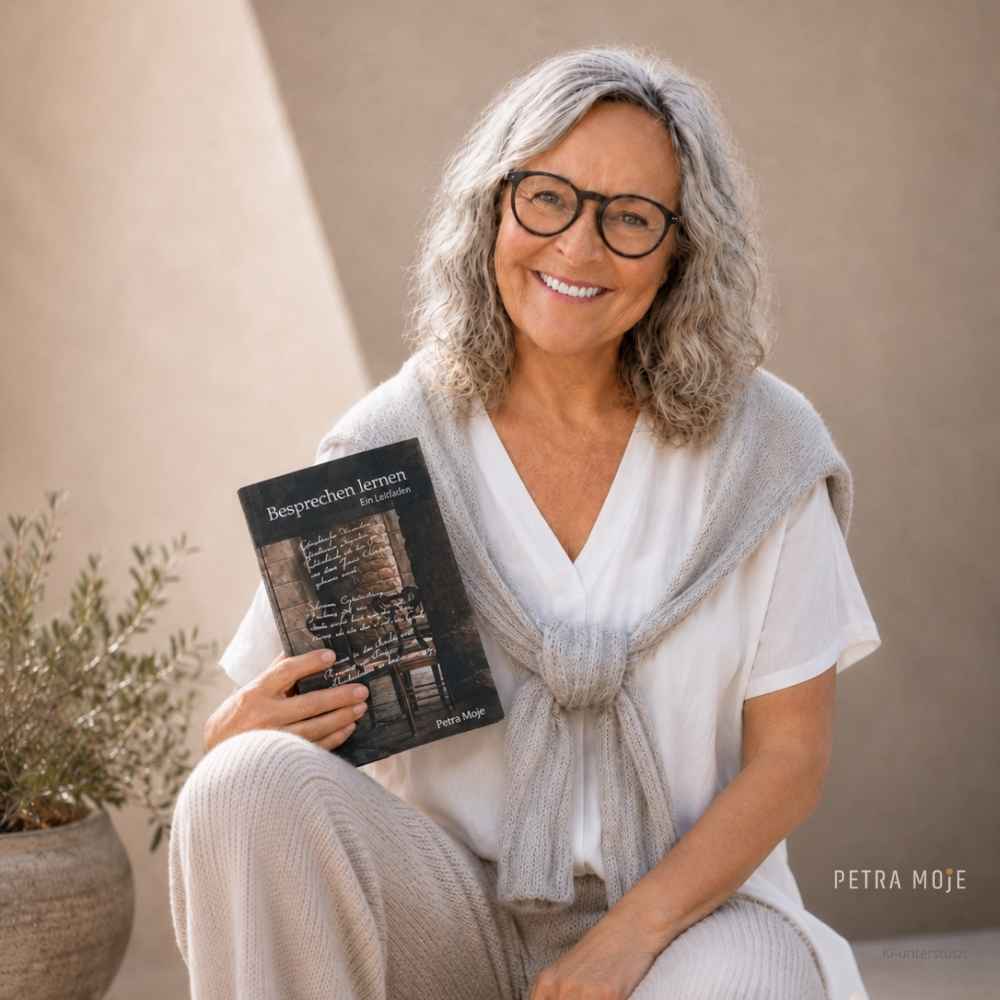 Petra Moje sitzt mit dem Buch „Besprechen lernen. Ein Leitfaden“ in der Hand und blickt lächelnd in die Kamera.